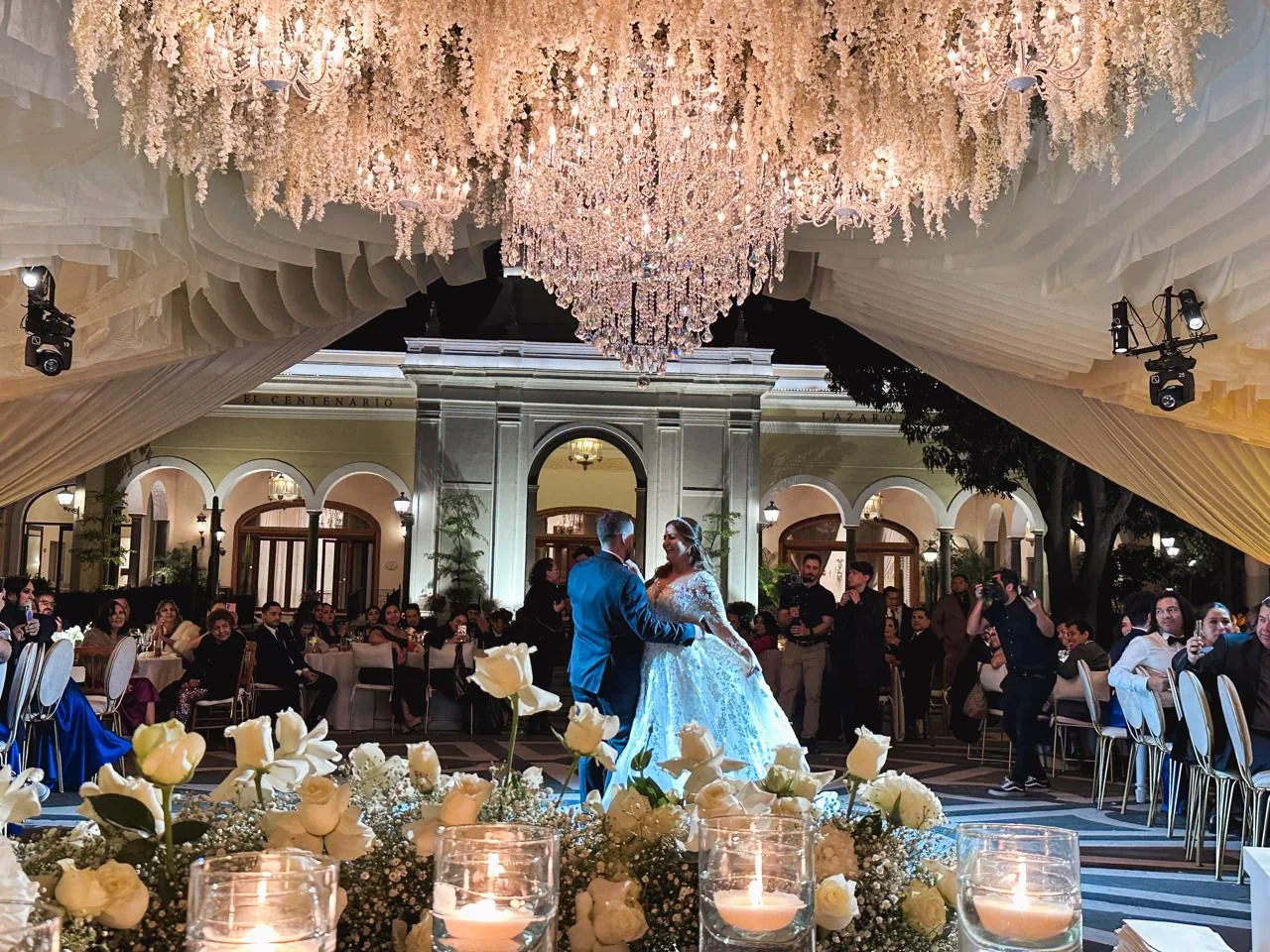 Pareja bailando bajo dosel entelado blanco con candelabros de cristal y arco floral en patio de hacienda