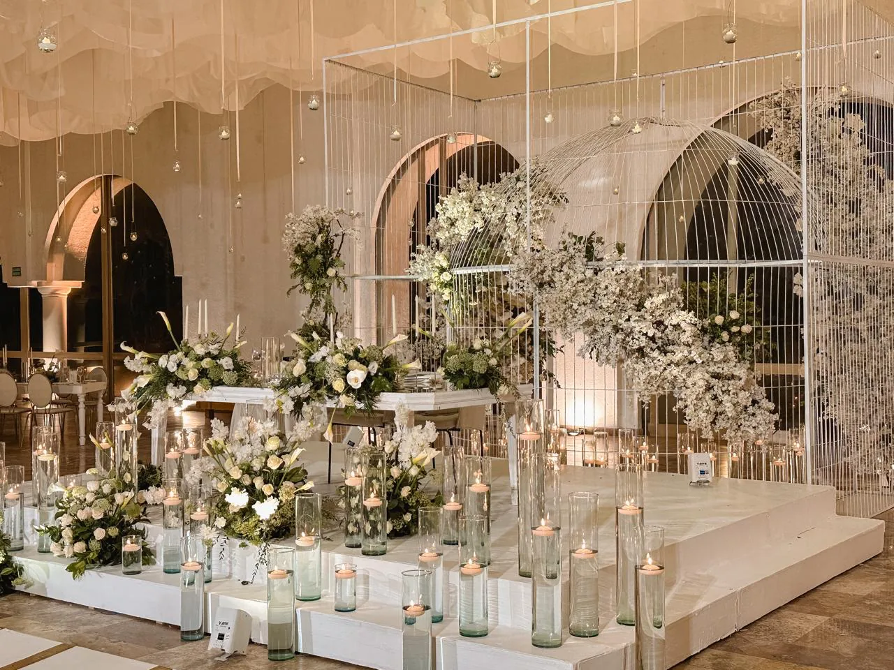 Escenario con jaula ornamental de alambre, arcos florales blancos y velas en pedestal