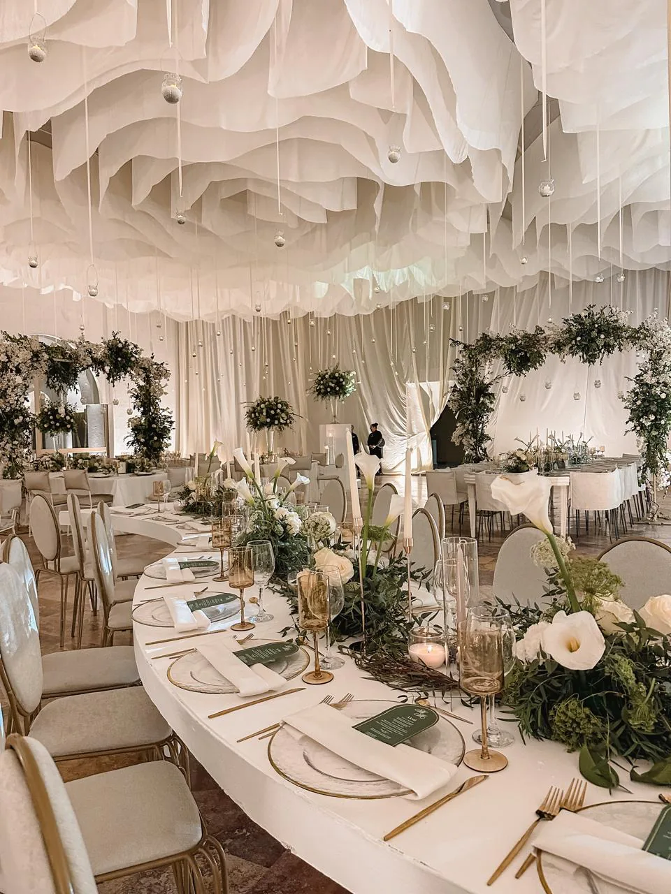 Mesa principal con techo entelado ondulado, arcos florales verdes y centro de mesa con calas